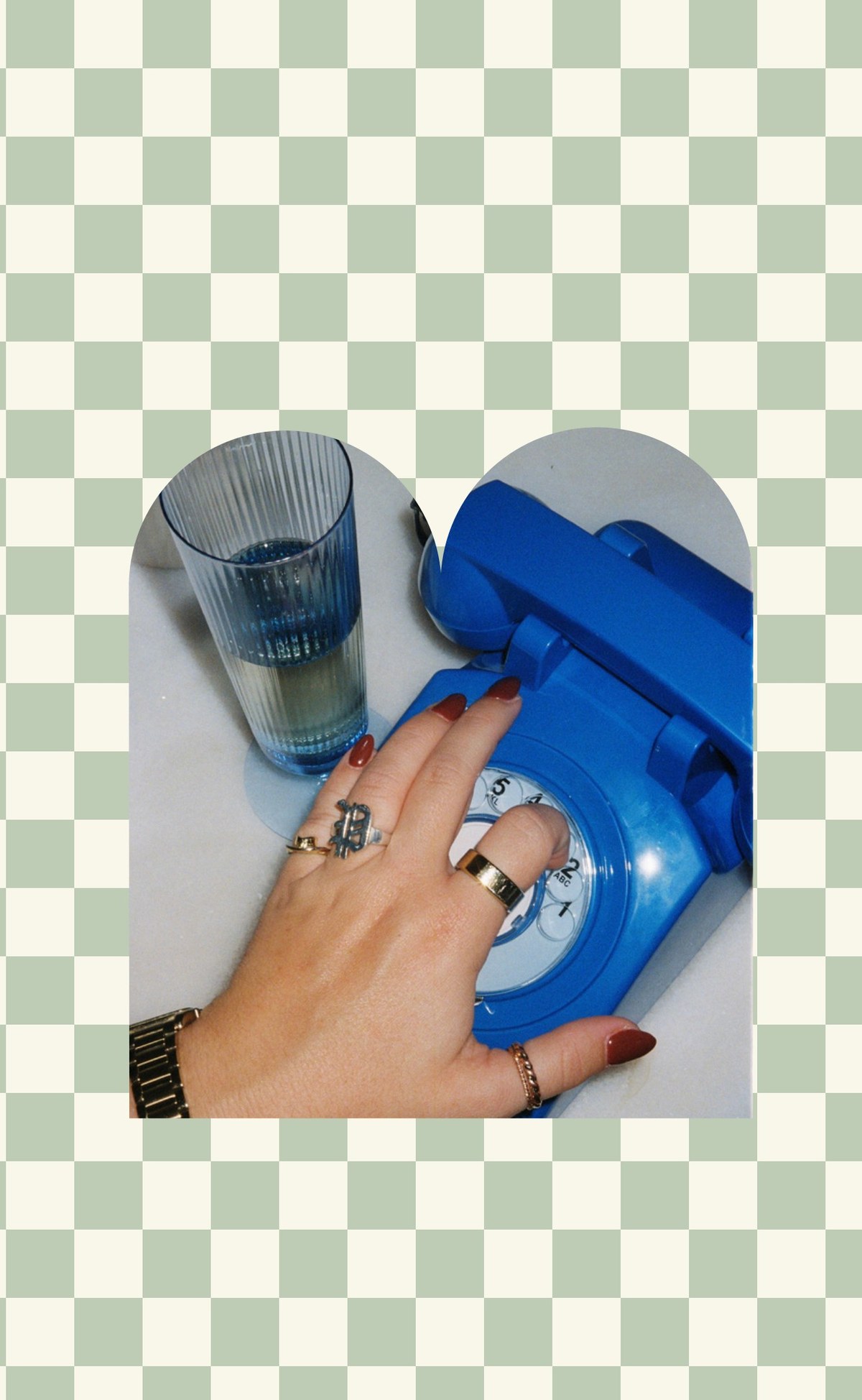 A hand with rings dialing a blue rotary phone next to a glass of water on a checkered background